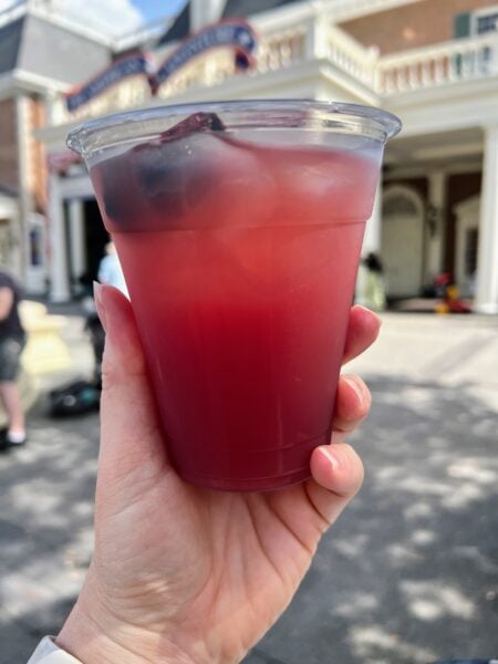 EPCOT Moonshine Sour over ice with a blackberry, held up in front of the American Adventure