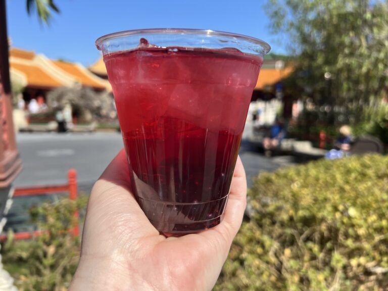 A hand holding a Honey Hibiscus Hard Iced Tea in a clear cup, with the China pavilion rooftops behind it at EPCOT