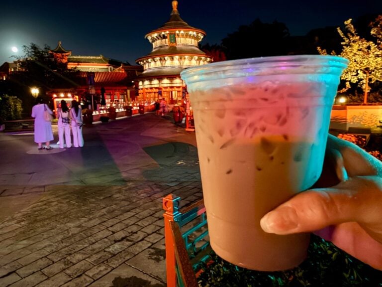 Hand holding a Tipsy Ducks in Love iced coffee cocktail in front of the Temple of Heaven at EPCOT's China pavilion at night, with the moon behind it